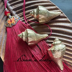May include: A decorative tassel with a red fringe, a floral design, and three gold-colored conch shell ornaments. The tassel is adorned with a red bead and a decorative floral charm. The conch shells are arranged on a wooden surface.