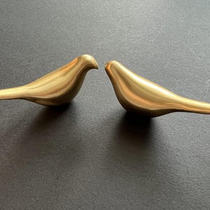 Pu&ograve; includere: Due figurine di uccelli in metallo dorato che si fronteggiano. Gli uccelli sono astratti e hanno una finitura liscia e lucida.