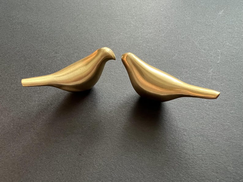Pu&ograve; includere: Due figurine di uccelli in metallo dorato che si fronteggiano. Gli uccelli sono astratti e hanno una finitura liscia e lucida.
