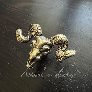 Puede incluir: Una escultura de calavera de carnero de latón con cuernos detallados y superficies texturizadas. La escultura es de color dorado y está sobre un fondo oscuro. La calavera de carnero es un objeto decorativo.