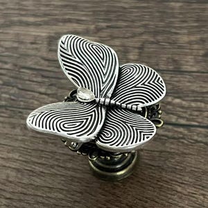 Può includere: Pomello per armadio a forma di farfalla color argento con una gemma trasparente al centro. Le ali della farfalla presentano un motivo dettagliato a forma di impronta digitale. Il pomello ha una base color bronzo ed è posto su una superficie di legno.