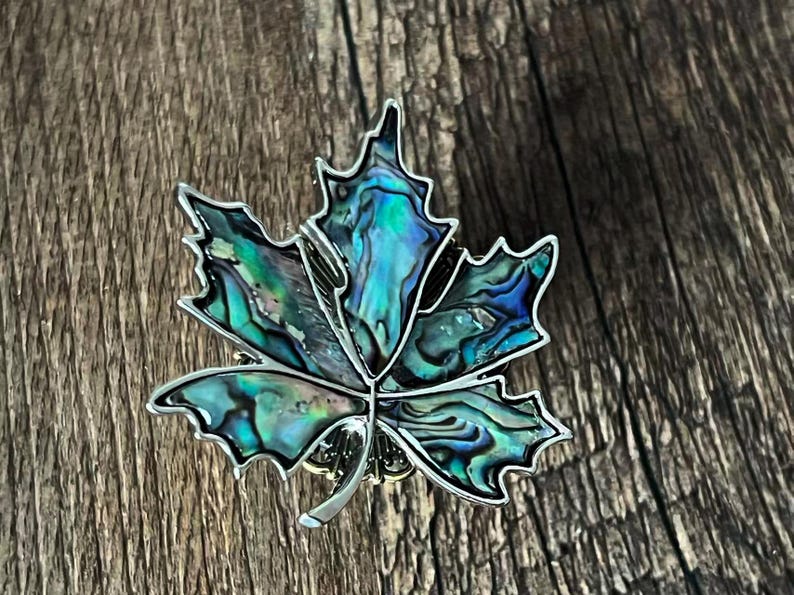 Peut inclure: Une broche en forme de feuille d'&eacute;rable argent&eacute;e avec une incrustation de coquillage d'ormeau. La coquille iris&eacute;e pr&eacute;sente des nuances de bleu, de vert et de violet. La broche est pos&eacute;e sur un fond en bois, soulignant la beaut&eacute; naturelle du design.