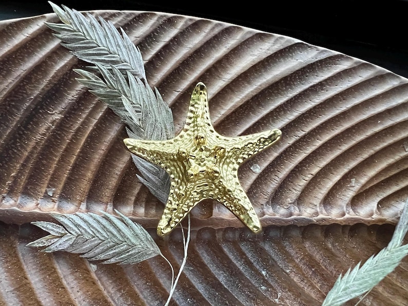 May include: A gold-coloured starfish ornament with a textured surface. The starfish is on a brown wooden background with dried leaves.
