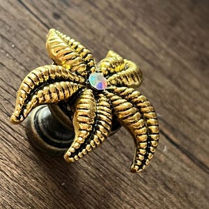 Op de afbeelding: Een decoratieve kastknop met een goudkleurig zeesterontwerp. De zeester heeft gedetailleerde ribbels en een centrale iriserende edelsteen. De knop is gemonteerd op een donkere, antieke basis, geschikt voor meubels of decoratie.