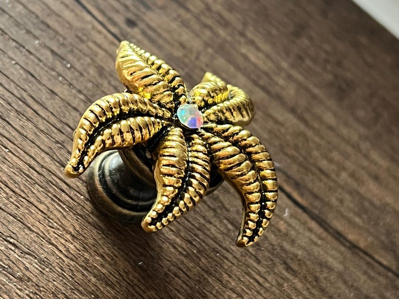 Op de afbeelding: Een decoratieve kastknop met een goudkleurig zeesterontwerp. De zeester heeft gedetailleerde ribbels en een centrale iriserende edelsteen. De knop is gemonteerd op een donkere, antieke basis, geschikt voor meubels of decoratie.