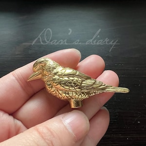 Puede incluir: Una figurita de pájaro de metal dorado con alas detalladas y un pico. El pájaro está posado en una mano.