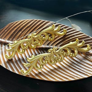 May include: Two ornate gold-colored drawer pulls with a decorative, floral design. The pulls are displayed on a wooden surface.