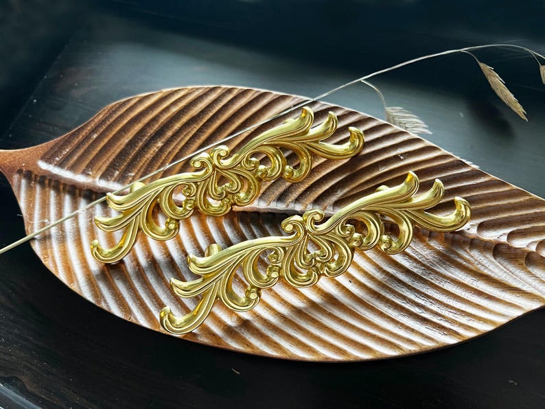 May include: Two ornate gold-colored drawer pulls with a decorative, floral design. The pulls are displayed on a wooden surface.