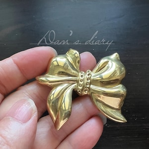 Pode incluir: Broche dourado em forma de laço. O broche tem um design detalhado com um centro texturizado e bordas lisas e curvas. O broche é segurado em uma mão contra um fundo escuro. O texto "Dan's diary" está escrito em itálico no fundo.