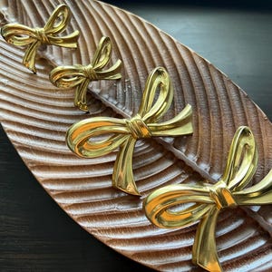 Può includere: Quattro oggetti decorativi dorati a forma di fiocco sono disposti su un vassoio di legno marrone a forma di foglia. I fiocchi hanno una finitura lucida e sono uniformemente distanziati sulla superficie del vassoio.