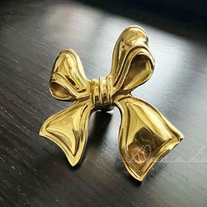 May include: A gold-coloured bow-shaped brooch with a polished, reflective surface. The bow has a detailed design with multiple folds and curves, creating a three-dimensional effect. The brooch is likely made of metal and is a decorative accessory.