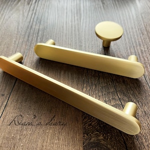 以下が含まれることがあります： 2つの長方形のプルと丸いノブを含む、金色のキャビネットハードウェアのセット。 ハードウェアはつや消し仕上げで、家具用に設計されています。 アイテムは木製の表面に表示されます。