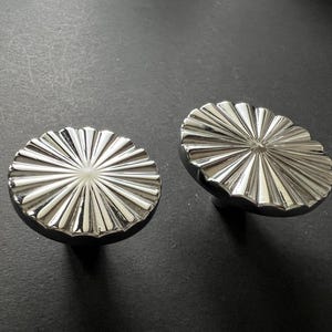 Peut inclure: Deux boutons de meuble argentés avec un motif radial. Les boutons ont une base sombre et une surface brillante et réfléchissante. Le design ressemble à un éclat de soleil ou à une fleur, ajoutant une touche décorative aux meubles.