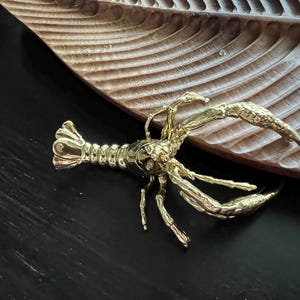 May include: A gold-coloured metal lobster figurine. The lobster is posed with its claws raised and its tail curled. The figurine is sitting on a wooden surface.