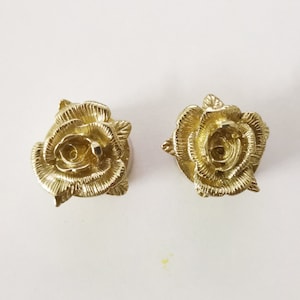 May include: Two gold-colored rose-shaped decorative embellishments. The roses are detailed with intricate carvings and have a slightly raised center.