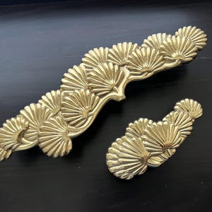 May include: Two gold-toned metal drawer pulls shaped like stylized pine branches. The larger pull has a curved shape and the smaller pull is straight.