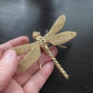 May include: A gold-coloured dragonfly figurine with detailed wing and body textures. The dragonfly has outstretched wings and is held against a dark background. The figurine is made of metal.