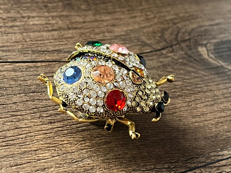 May include: A gold-coloured ladybird brooch encrusted with clear and colourful gemstones. The brooch features a detailed design with a mix of red, blue, pink, and orange stones, and measures approximately 4 cm in length.