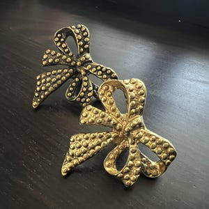 Pode incluir: Duas broches douradas em forma de laço com uma superfície texturizada e pontilhada. As broches estão posicionadas sobre uma superfície de madeira escura. Os laços têm um design clássico, com laços e caudas.