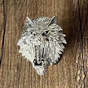 K&ouml;nnte beinhalten: Silberfarbener Wolfskopf-Anh&auml;nger mit offenem Maul, das scharfe Z&auml;hne zeigt. Der Anh&auml;nger hat eine detaillierte Fellstruktur und ist vor einem Holzgrund platziert. Die Augen des Wolfes sind dunkel.