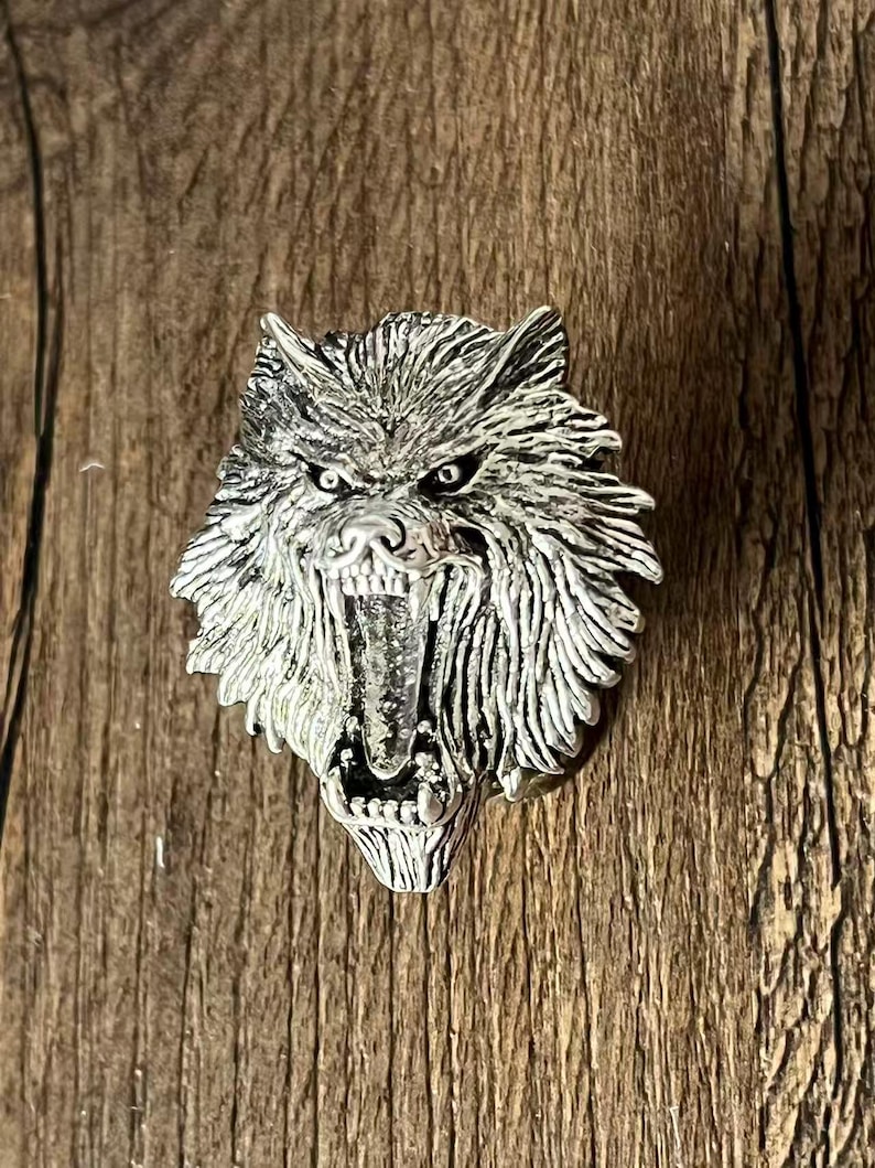 K&ouml;nnte beinhalten: Silberfarbener Wolfskopf-Anh&auml;nger mit offenem Maul, das scharfe Z&auml;hne zeigt. Der Anh&auml;nger hat eine detaillierte Fellstruktur und ist vor einem Holzgrund platziert. Die Augen des Wolfes sind dunkel.