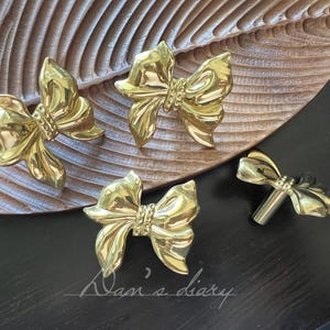 May include: Four gold-colored bow-shaped cabinet knobs. The knobs are arranged on a wooden leaf-shaped tray and a dark surface. The bows have detailed textures and a shiny finish. The image includes the text "Dan's diary".