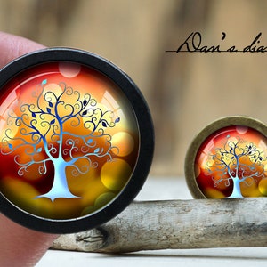 May include: A round, black framed glass cabochon with a colorful image of a stylized tree with blue and black branches and a white trunk. The background is a gradient of orange, yellow, and red with a blurred pattern of circles. The image is framed by a black metal ring.
