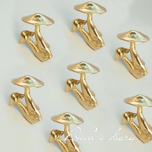 Op de afbeelding: Acht goudkleurige paddenstoelvormige sculpturen met een glad, getextureerd oppervlak. Elk beeldhouwwerk heeft een afgeronde bovenkant en een steel met een unieke, organische vorm.