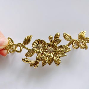 Puede incluir: Diseño floral de metal dorado con dos flores y hojas. El diseño es intrincado y detallado.