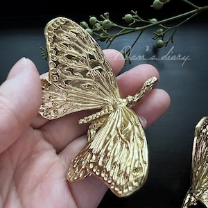 Op de afbeelding: Een gedetailleerd, goudkleurig vlinderornament met ingewikkelde vleugelpatronen. De vlinder wordt in een hand gehouden, wat het delicate ontwerp laat zien. De afbeelding staat tegen een donkere achtergrond, wat de metaalglans van de vlinder benadrukt.