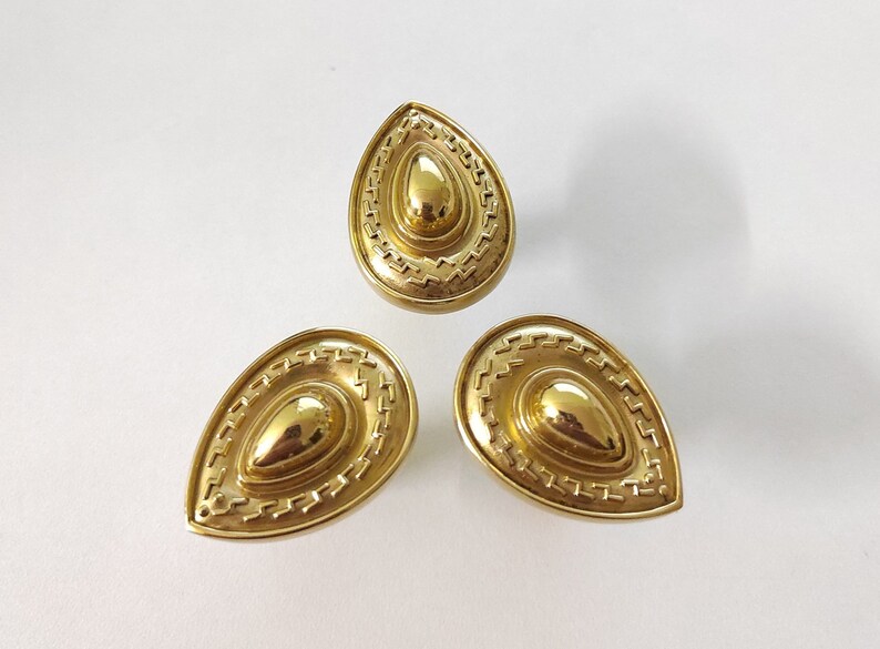 May include: Three gold-toned teardrop-shaped cabinet knobs with a decorative, concentric circle design.
