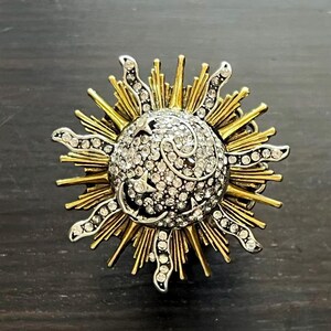 May include: A sun-shaped brooch with a central, crystal-studded sphere. The brooch features gold-colored rays and silver-colored accents. The sun design is a detailed, decorative accessory.