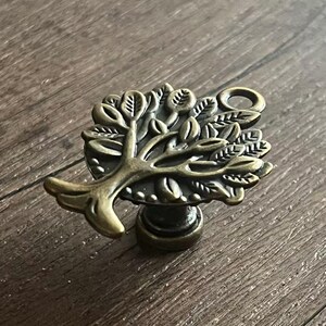 Op de afbeelding: Een antieke bronzen kastknop met een boomontwerp. De knop heeft een gedetailleerde boom met bladeren en takken, en een kleine lus voor bevestiging. De knop is gemonteerd op een donkerbruin houten oppervlak.