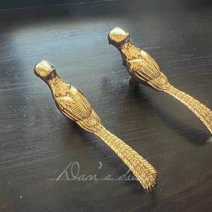 Op de afbeelding: Twee goudkleurige vogelvormige lade- of kastgrepen. Het lichaam van de vogel is gedetailleerd met geëtste veren en een lange staart. De handgrepen zijn geplaatst op een donkere houten ondergrond.