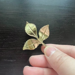 May include: A gold-coloured metal leaf sprig with three leaves. The leaves are detailed with veins and have a slightly textured surface.