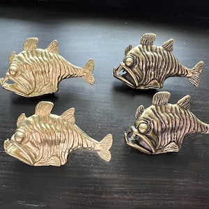 May include: Four gold-coloured metal fish sculptures with detailed features. The fish have a textured, scaly appearance and are positioned on a dark surface.