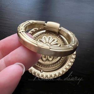 May include: A gold-toned metal ring pull with a decorative floral design. The ring pull is attached to a round, decorative base.