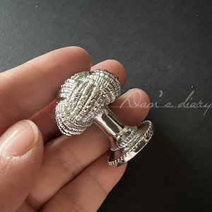 Könnte beinhalten: Silberner Schrank-Knopf mit einem dekorativen Knotendesign. Der Knopf wird in einer Hand gehalten.