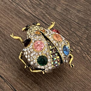 May include: A gold-coloured ladybird brooch, adorned with clear and colourful rhinestones. The brooch has a detailed design with various coloured gemstones, including pink, green, blue, and orange. The ladybird's body is covered in sparkling crystals.