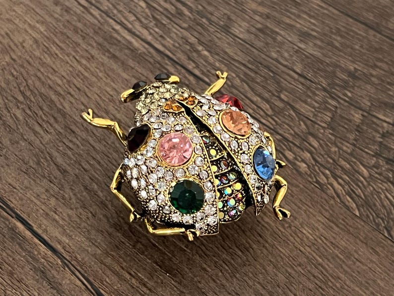 May include: A gold-coloured ladybird brooch, adorned with clear and colourful rhinestones. The brooch has a detailed design with various coloured gemstones, including pink, green, blue, and orange. The ladybird's body is covered in sparkling crystals.