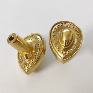 May include: Two gold-toned cabinet knobs with a decorative, ornate design. The knobs are shaped like a teardrop with a raised, rounded center.