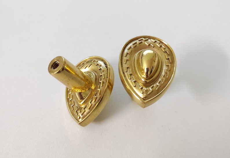 May include: Two gold-toned cabinet knobs with a decorative, ornate design. The knobs are shaped like a teardrop with a raised, rounded center.