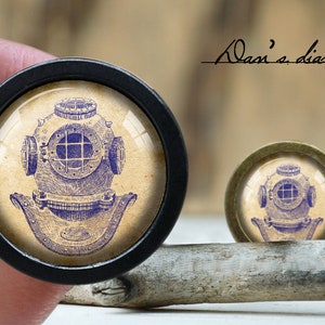 Puede incluir: Dos pomos redondos para armario con un diseño de casco de buzo vintage. El pomo más grande tiene un marco negro, el más pequeño un marco bronce. La ilustración del casco de buzo es azul sobre fondo beige.
