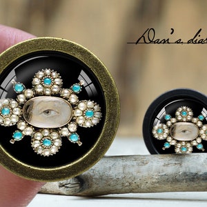 May include: Two decorative cabinet knobs with a vintage design. Each knob features a black background with an eye-shaped image surrounded by white pearls and turquoise accents. The larger knob has a bronze-colored frame, and the smaller one has a black frame. The text "Dan's diary" is visible.