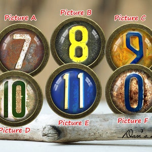 Pode incluir: Seis cabochões redondos, de metal, decorativos, cobertos de vidro, com os números 7, 8, 9, 10, 11 e 0. Os números são pintados em branco, amarelo e azul sobre um fundo marrom, amarelo e azul. Os cabochões estão dispostos em duas filas de três. Os cabochões são etiquetados com as letras A a F.