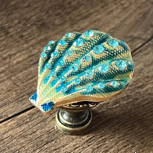 Può includere: Pomello decorativo per mobili con un design a conchiglia. Il pomello presenta una combinazione di colori turchese e oro, con strass turchesi. Il pomello è montato su una base color bronzo. Il design a conchiglia è dettagliato e accattivante.