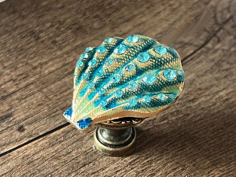 May include: A decorative cabinet knob with a seashell design. The knob has a teal and gold colour scheme, with teal-coloured rhinestones. The knob is mounted on a bronze-coloured base. The seashell design is detailed and eye-catching.
