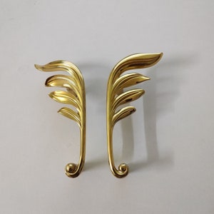 May include: Two gold-toned metal decorative door handles with a leaf design. The handles are shaped like a stylized leaf with a rounded bottom.