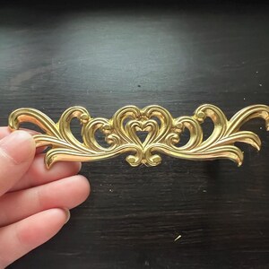 May include: A gold-colored metal decorative trim with a swirling, ornate design. The trim features a central heart shape with two smaller hearts on either side.