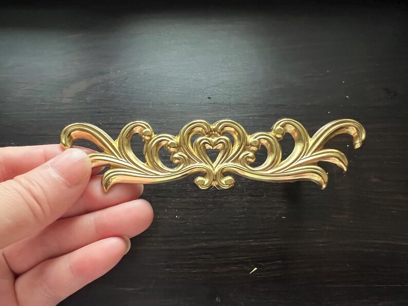 May include: A gold-colored metal decorative trim with a swirling, ornate design. The trim features a central heart shape with two smaller hearts on either side.